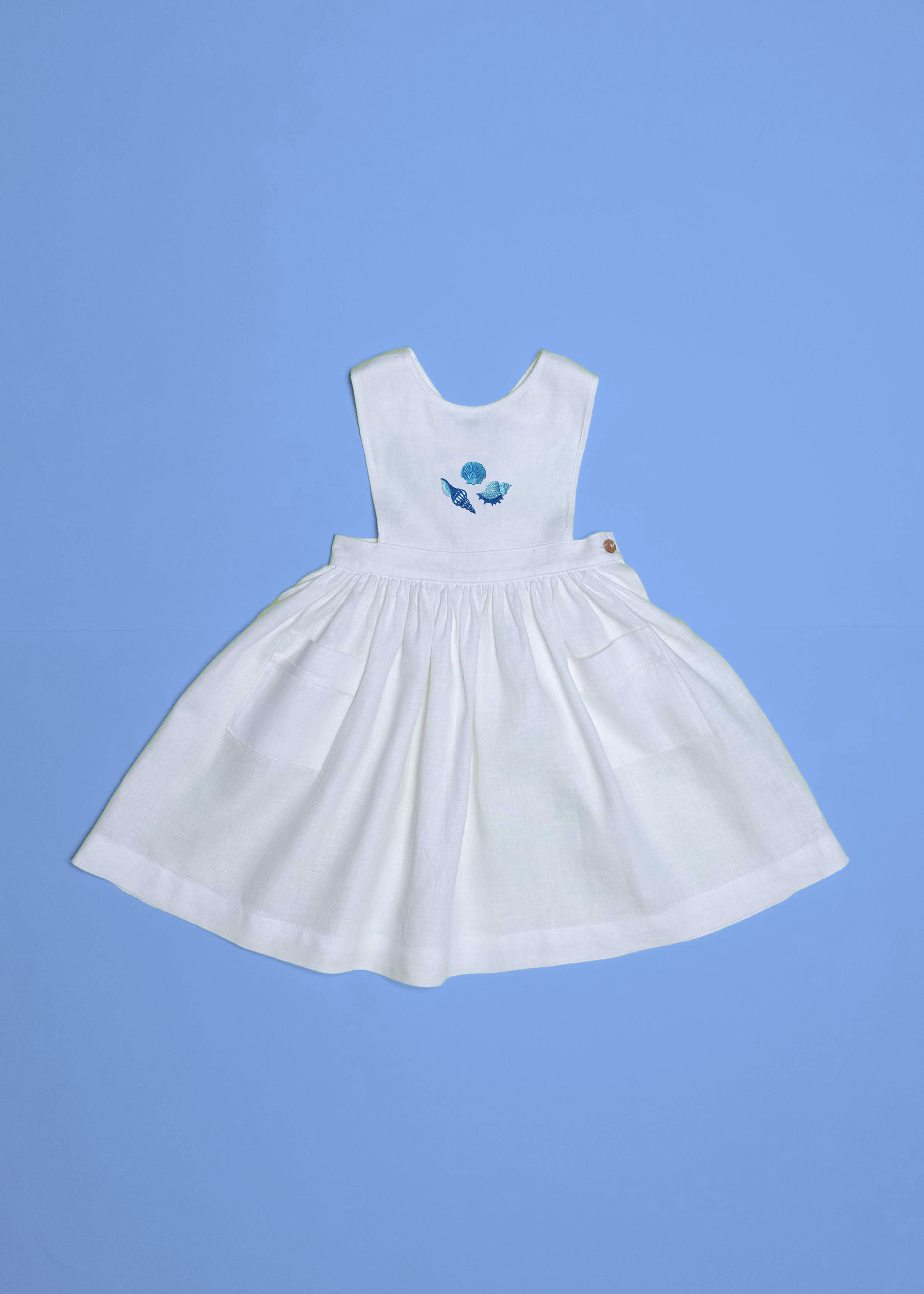 Vestido infantil de linho branco com bordado à mão de conchas azuis para meninas, visto de frente em superfície azul.