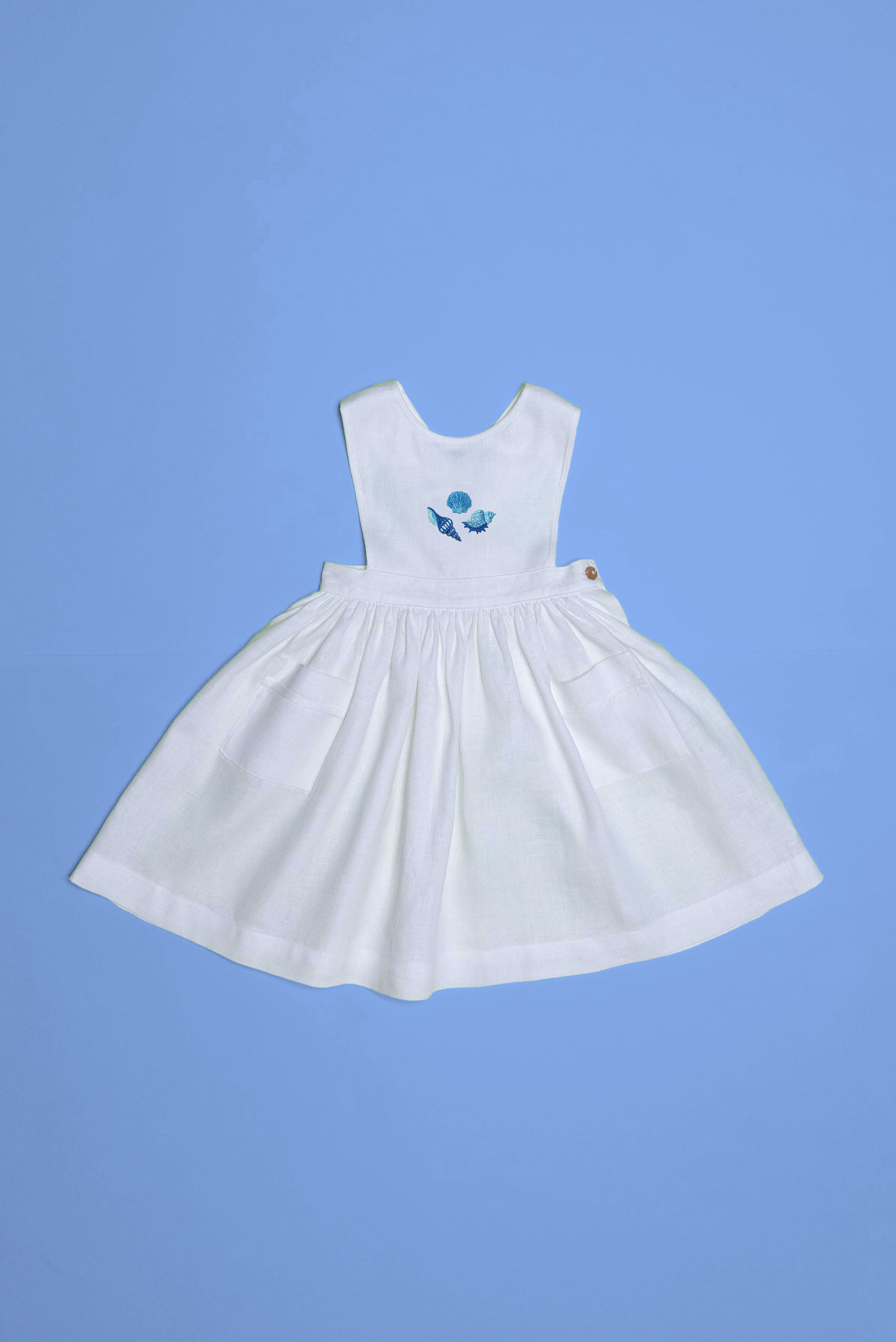 Vestido infantil de linho branco com bordado à mão de conchas azuis para meninas, visto de frente em superfície azul.