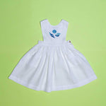 Vestido infantil de linho branco com bordado à mão de conchas azuis para meninas, visto de frente em superfície verde.