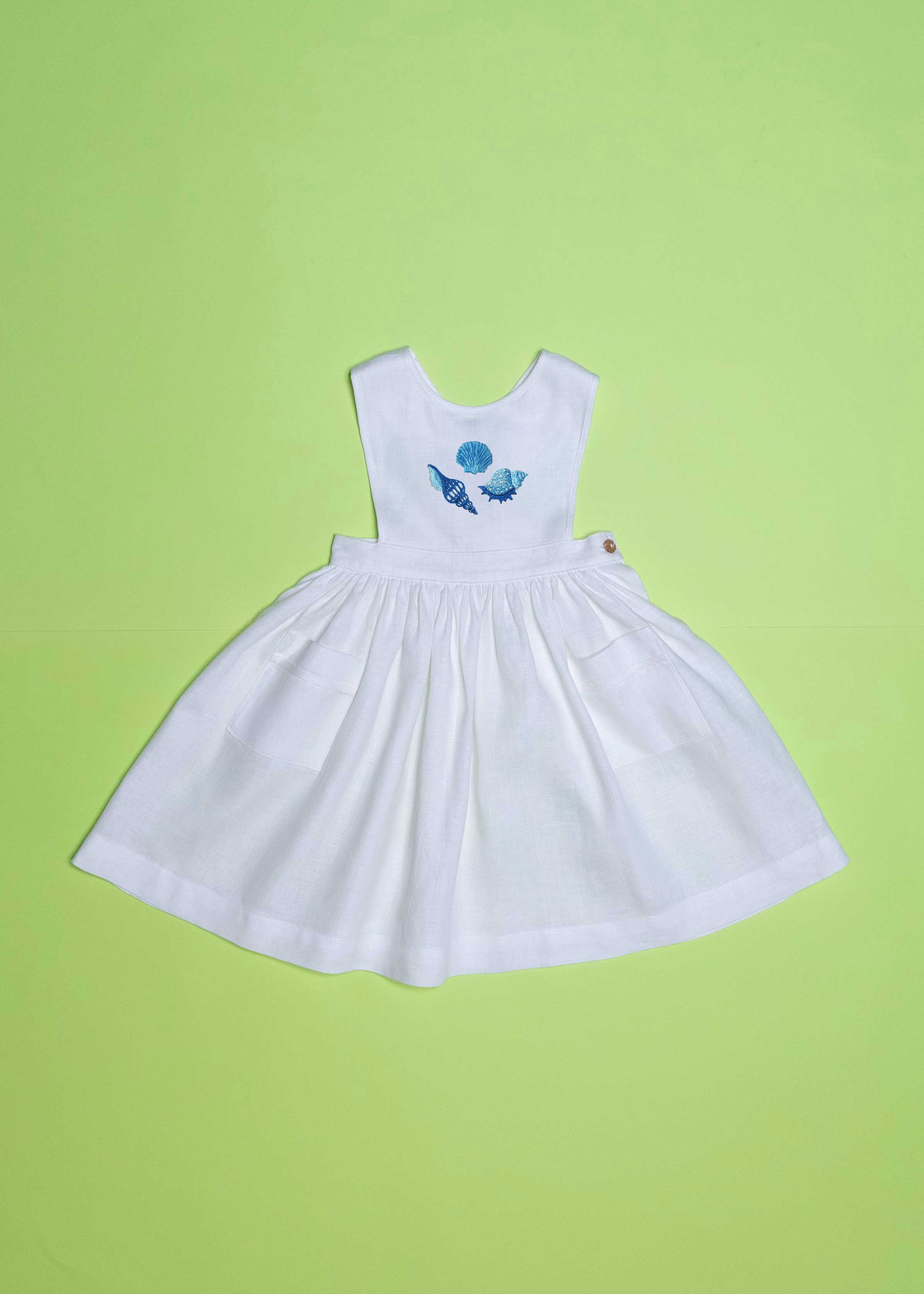 Vestido infantil de linho branco com bordado à mão de conchas azuis para meninas, visto de frente em superfície verde.