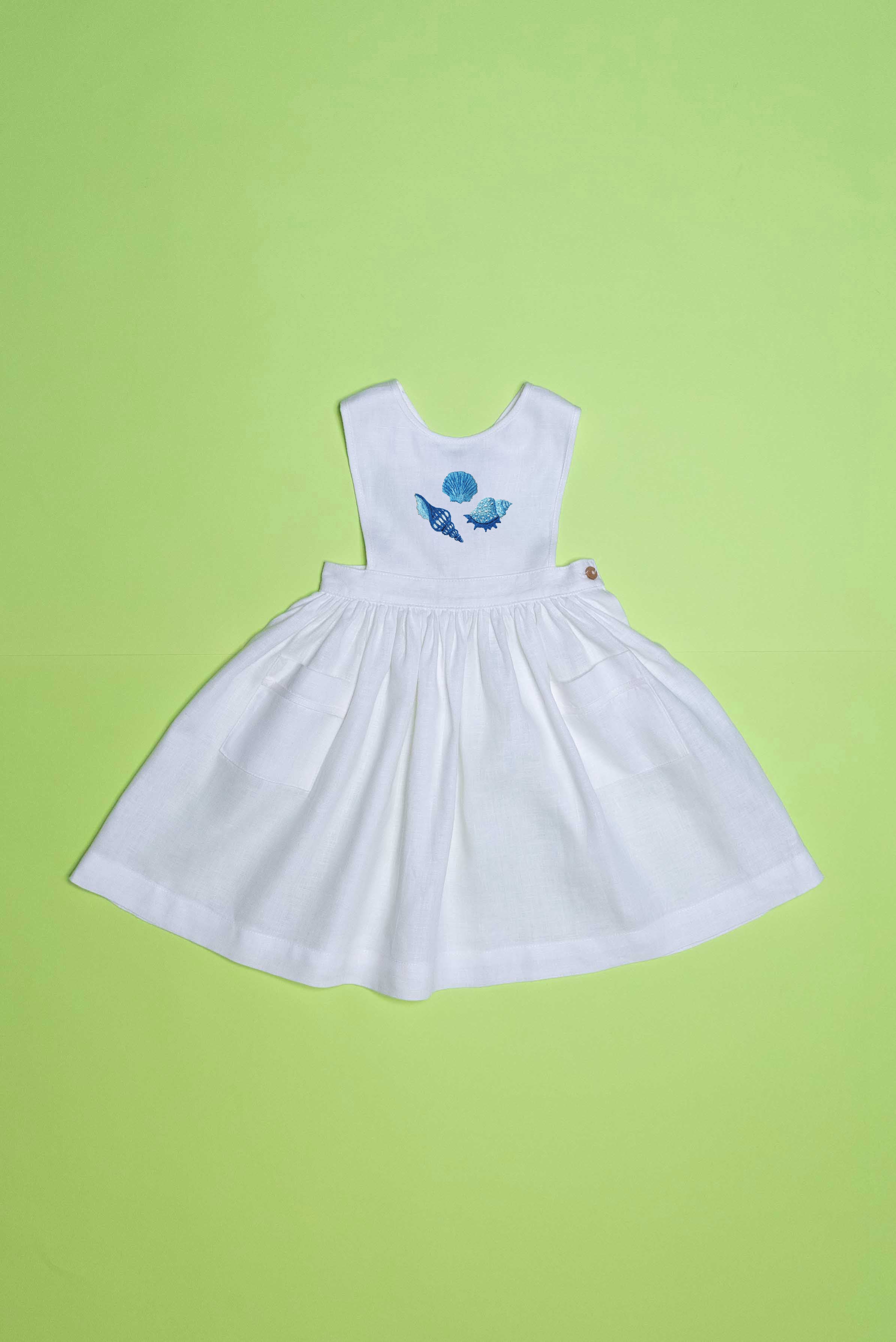 Vestido infantil de linho branco com bordado à mão de conchas azuis para meninas, visto de frente em superfície verde.