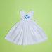 Vestido infantil de linho branco com bordado à mão de conchas azuis para meninas, visto de frente em superfície verde.