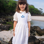 Menina na praia usando vestido infantil de linho branco com saia ampla pregueada, bolsos laterais e bordado de conchas do mar em tom azul na pala.