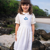 Menina na praia usando vestido infantil de linho branco com saia ampla pregueada, bolsos laterais e bordado de conchas do mar em tom azul na pala.