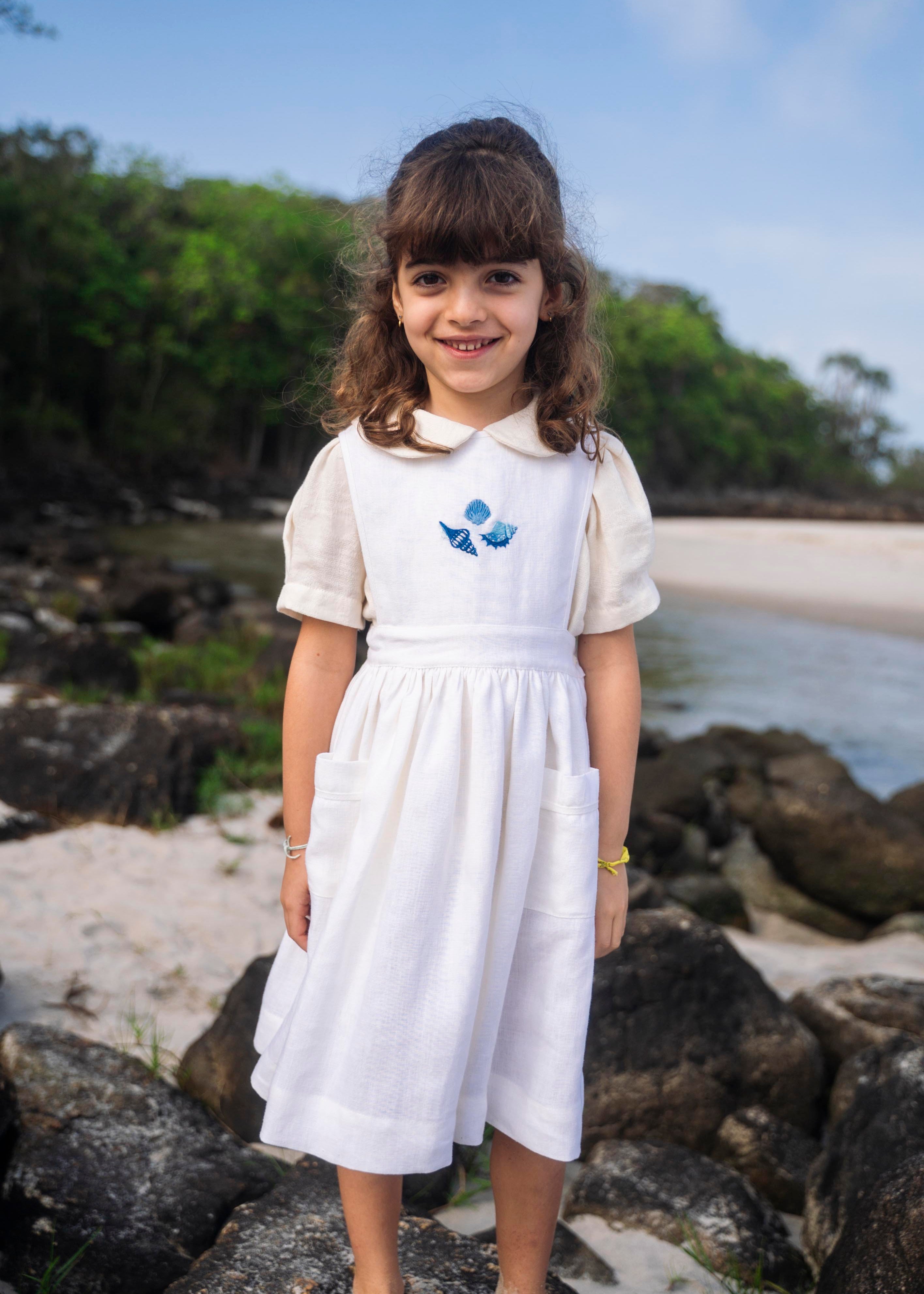 Menina na praia usando vestido infantil de linho branco com saia ampla pregueada, bolsos laterais e bordado de conchas do mar em tom azul na pala.