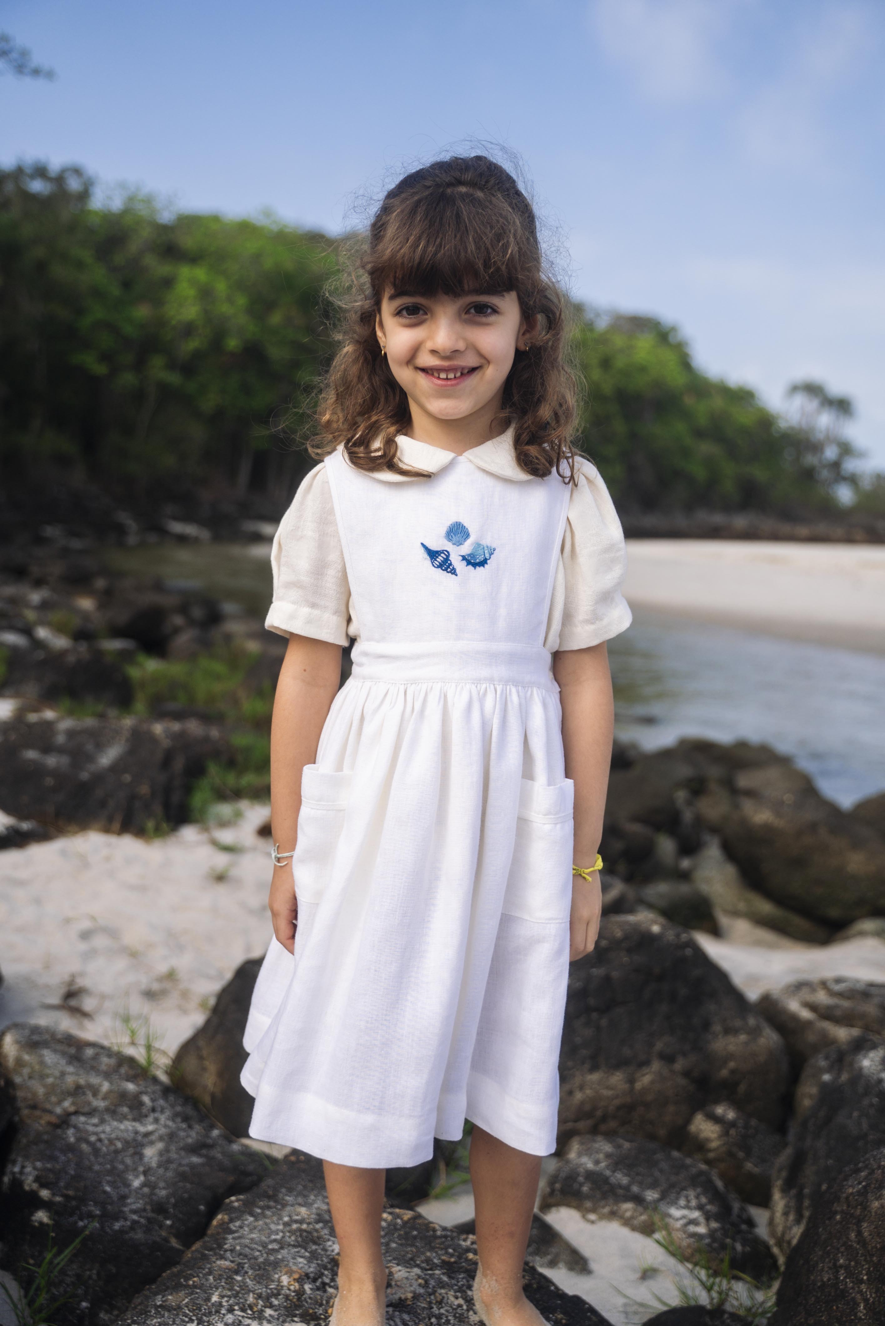 Menina na praia usando vestido infantil de linho branco com saia ampla pregueada, bolsos laterais e bordado de conchas do mar em tom azul na pala.