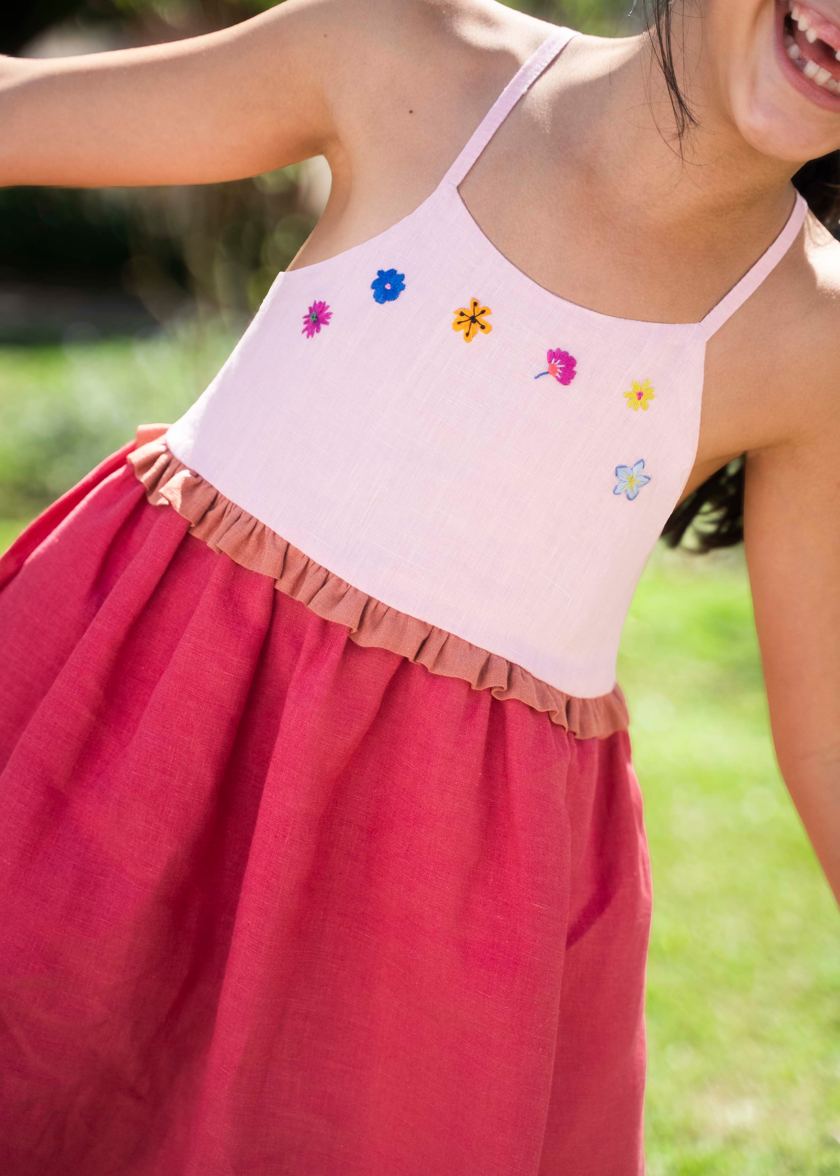 Menina usando vestido infantil em linho cereja e rosa com bordado de flores coloridas e franzido na cintura.