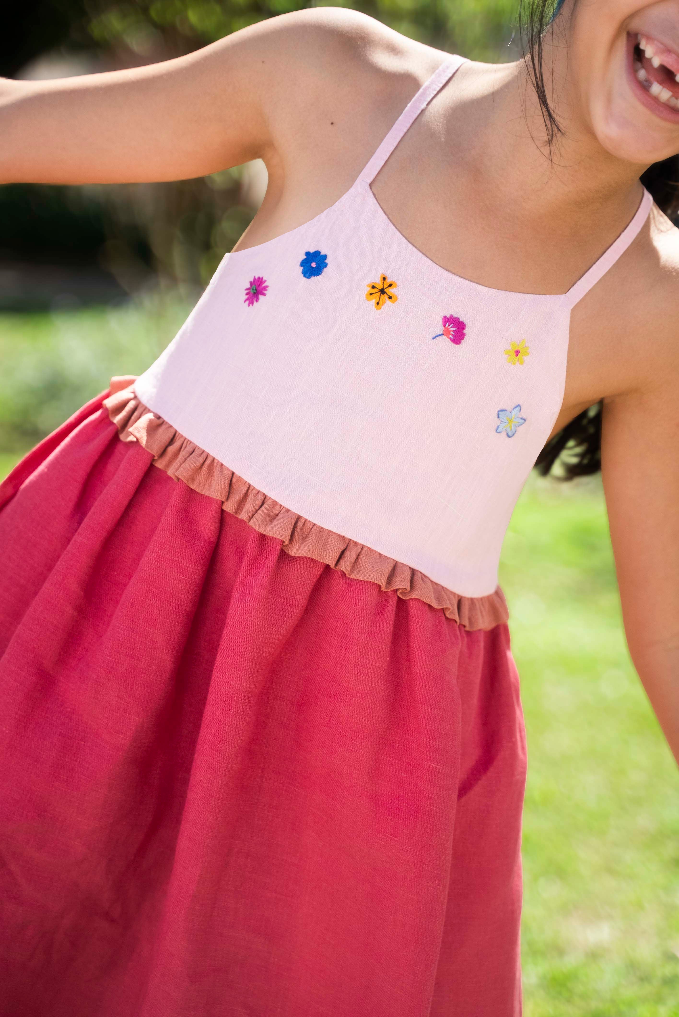 Menina usando vestido infantil em linho cereja e rosa com bordado de flores coloridas e franzido na cintura.