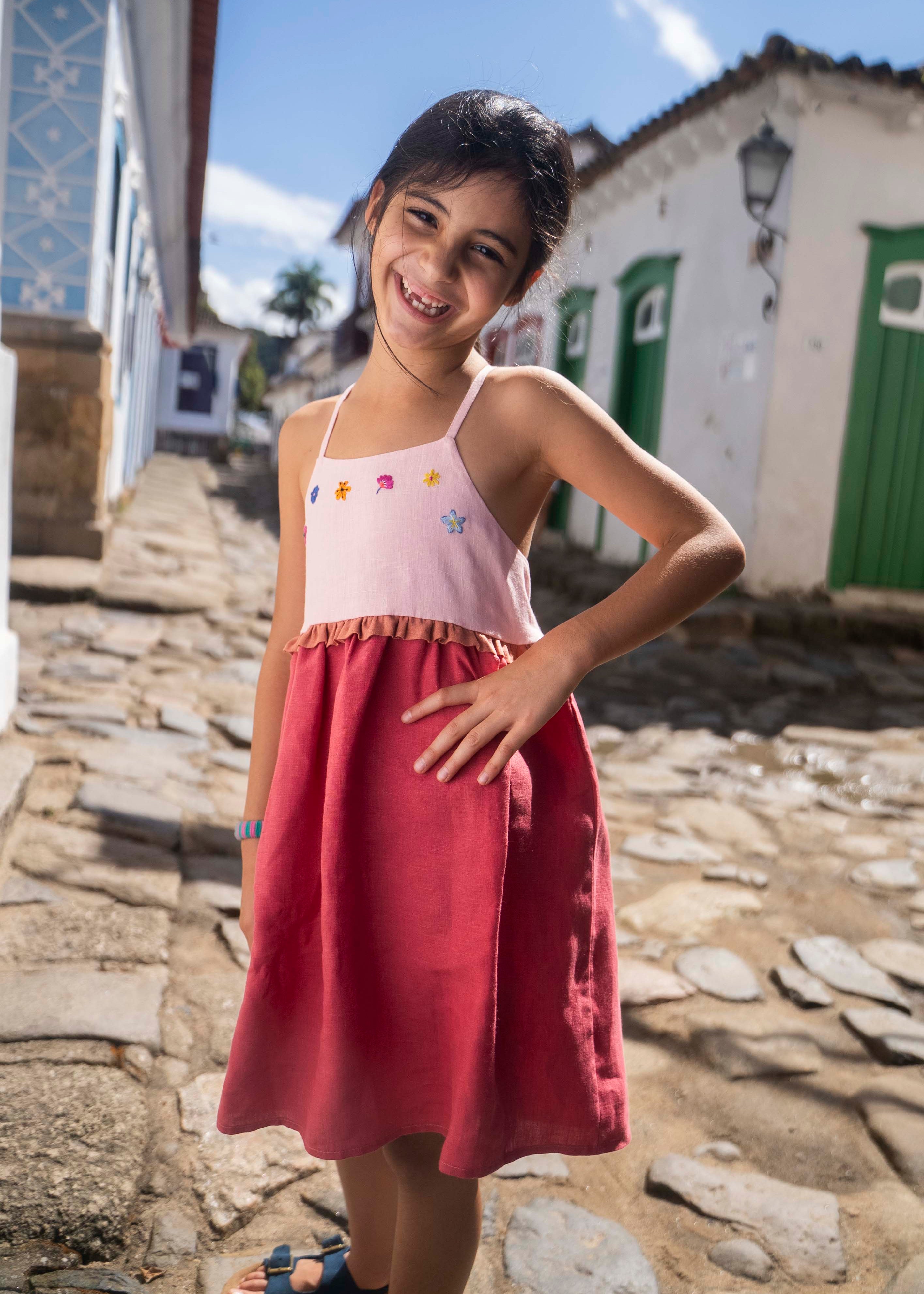 Menina usando vestido infantil em linho cereja e rosa com bordado de flores coloridas.