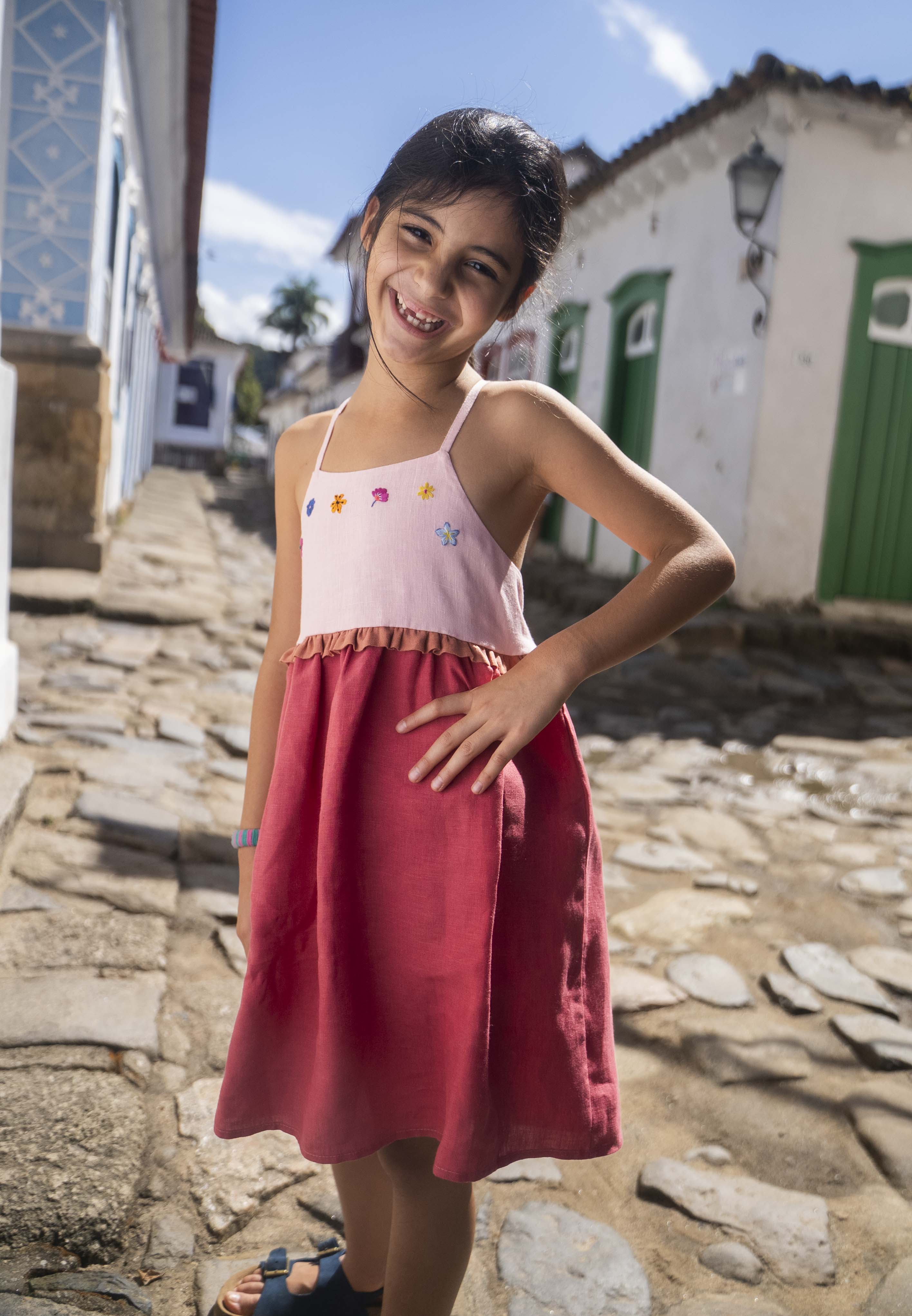 Menina usando vestido infantil em linho cereja e rosa com bordado de flores coloridas.