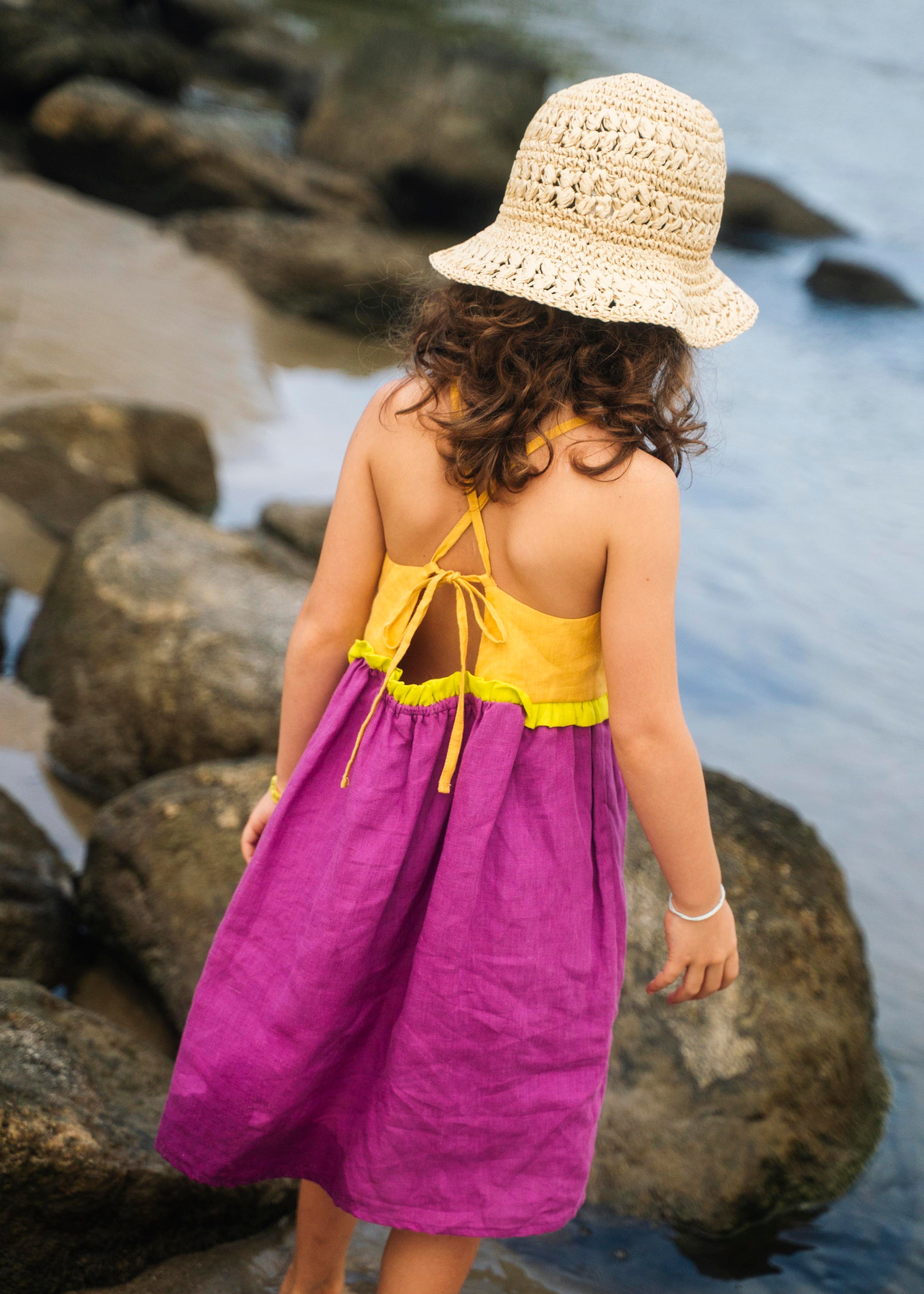 Costas do vestido infantil em linho roxo e amarelo com amarração ajustável e detalhe franzido verde, em look de verão.