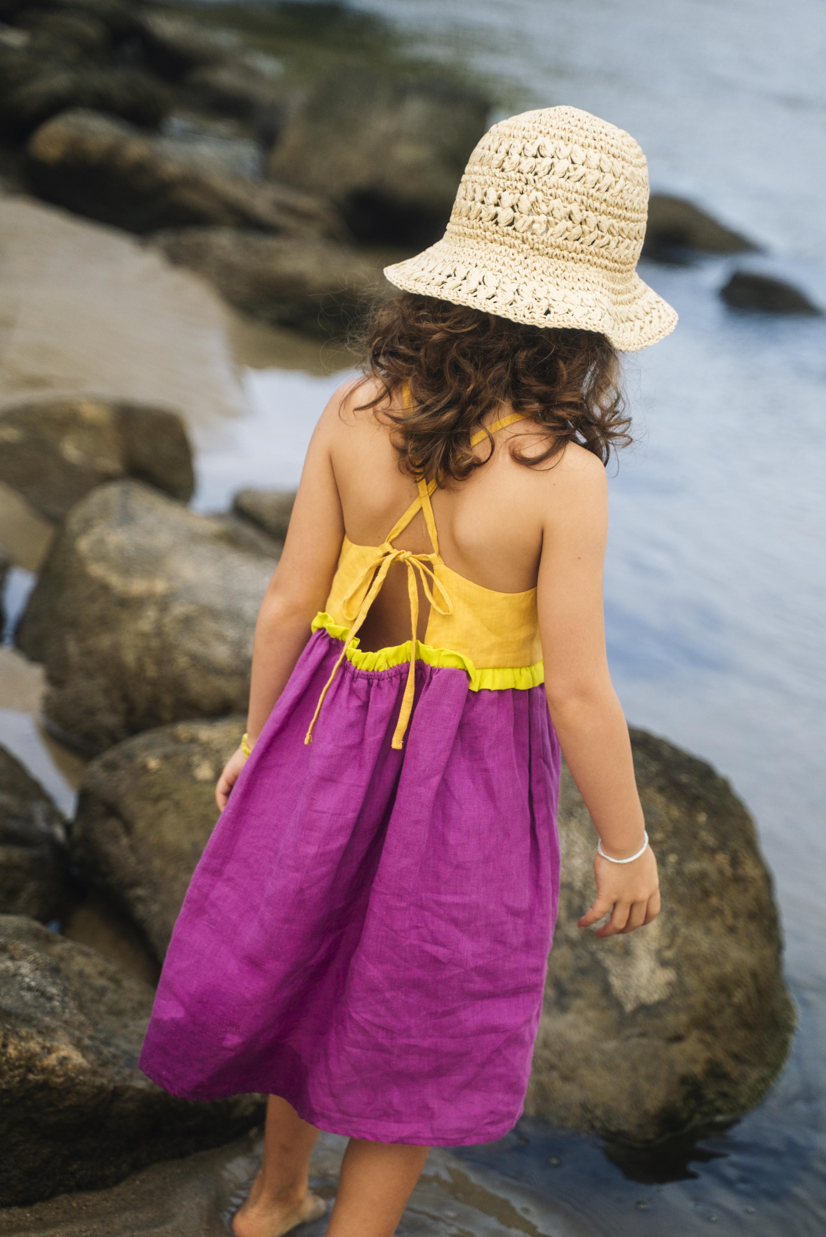 Costas do vestido infantil em linho roxo e amarelo com amarração ajustável e detalhe franzido verde, em look de verão.