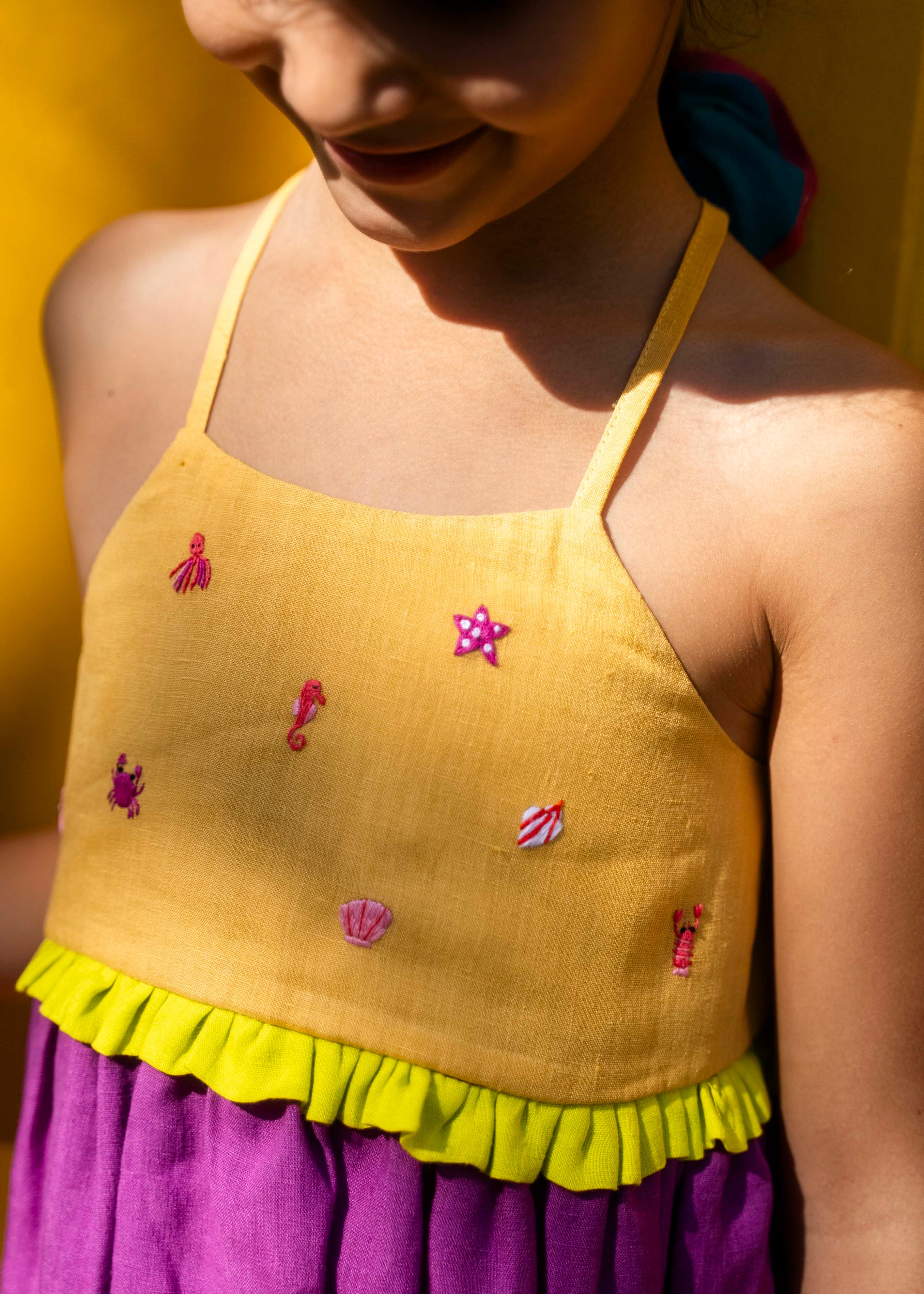 Close de menina com vestido infantil em linho roxo com alças finas e pala amarela com mini bordados manuais de bichinhos do fundo do mar, como polvo e cavalo-marinho.
