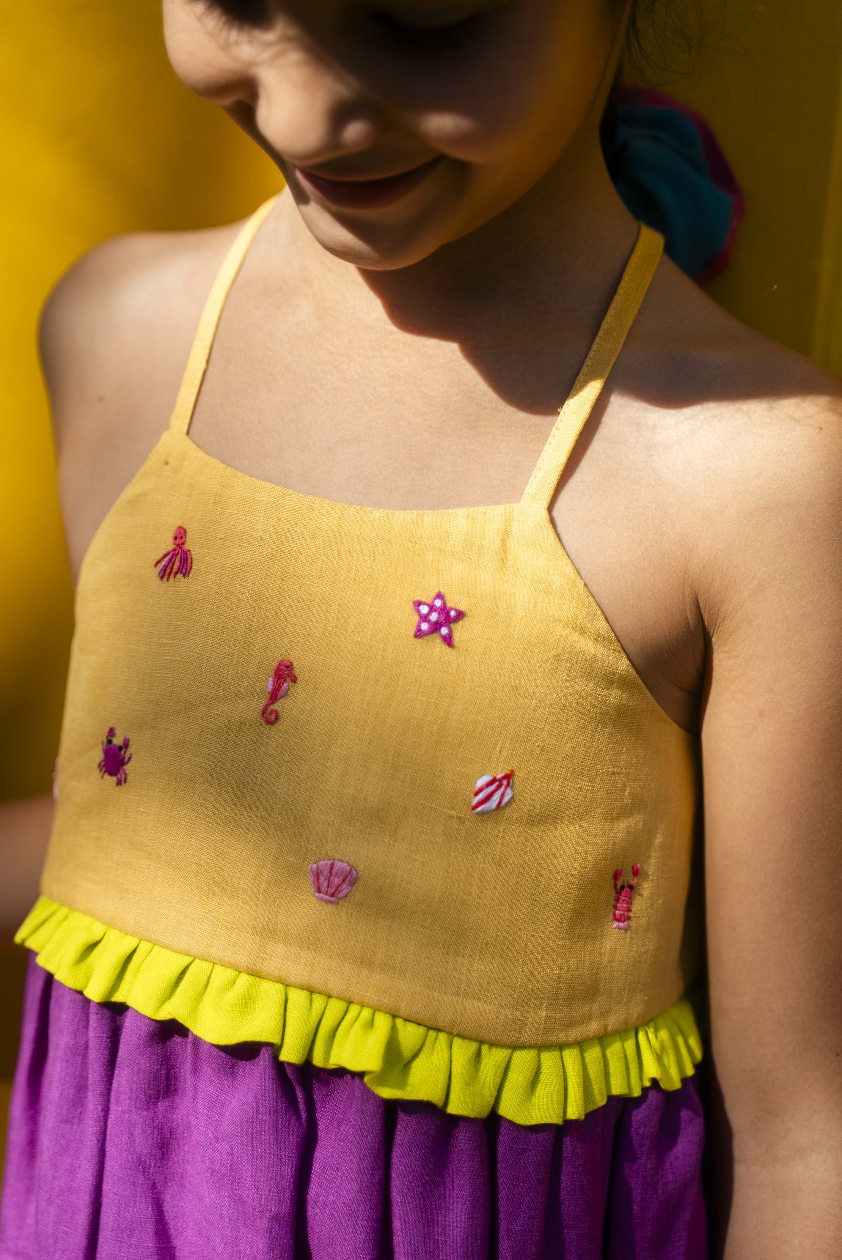Close de menina com vestido infantil em linho roxo com alças finas e pala amarela com mini bordados manuais de bichinhos do fundo do mar, como polvo e cavalo-marinho.