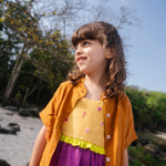 Menina usando vestido infantil em linho roxo e amarelo com bordados manuais de conchas, polvo e cavalo-marinho, com camisa marrom sobreposta.
