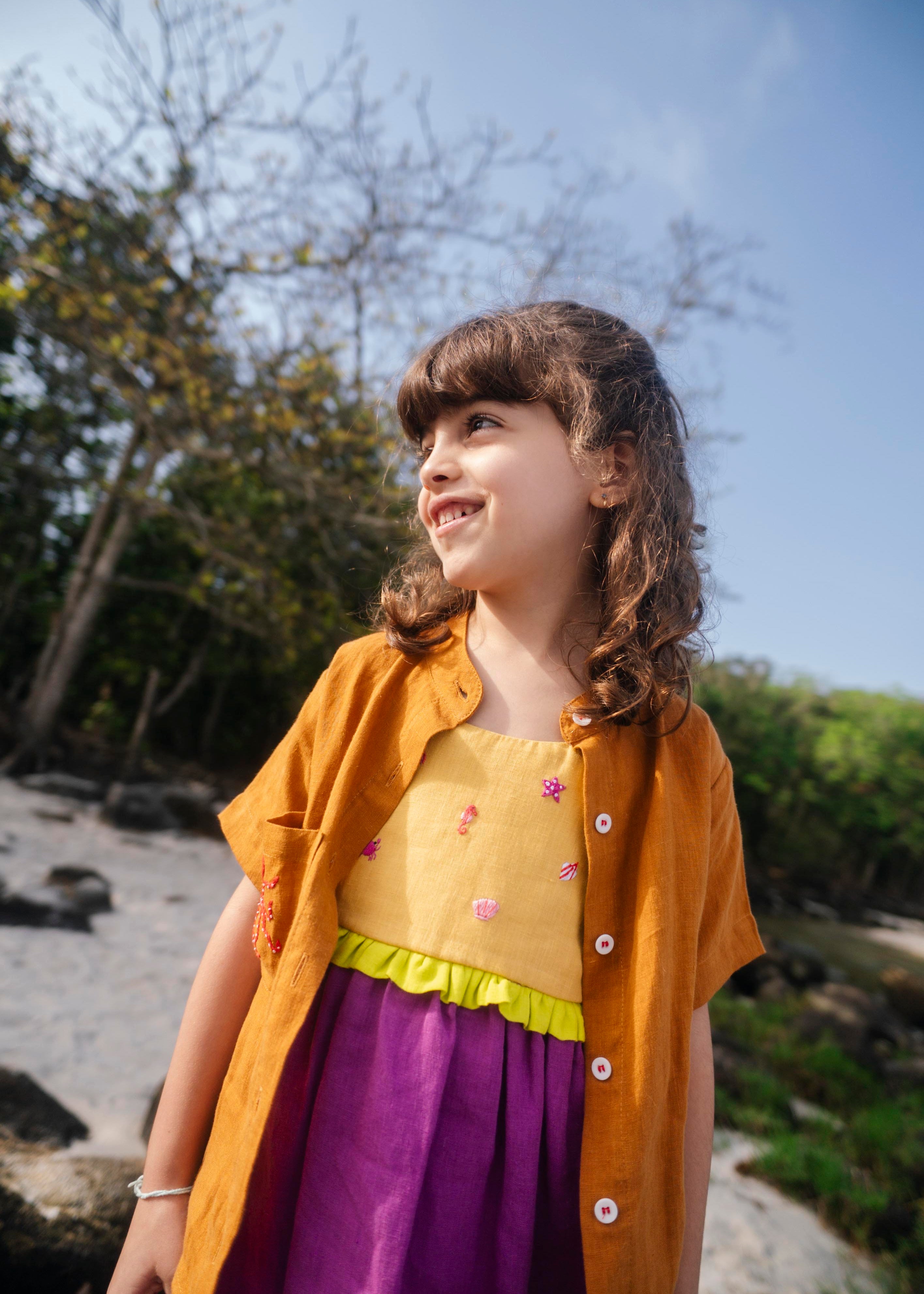 Menina usando vestido infantil em linho roxo e amarelo com bordados manuais de conchas, polvo e cavalo-marinho, com camisa marrom sobreposta.