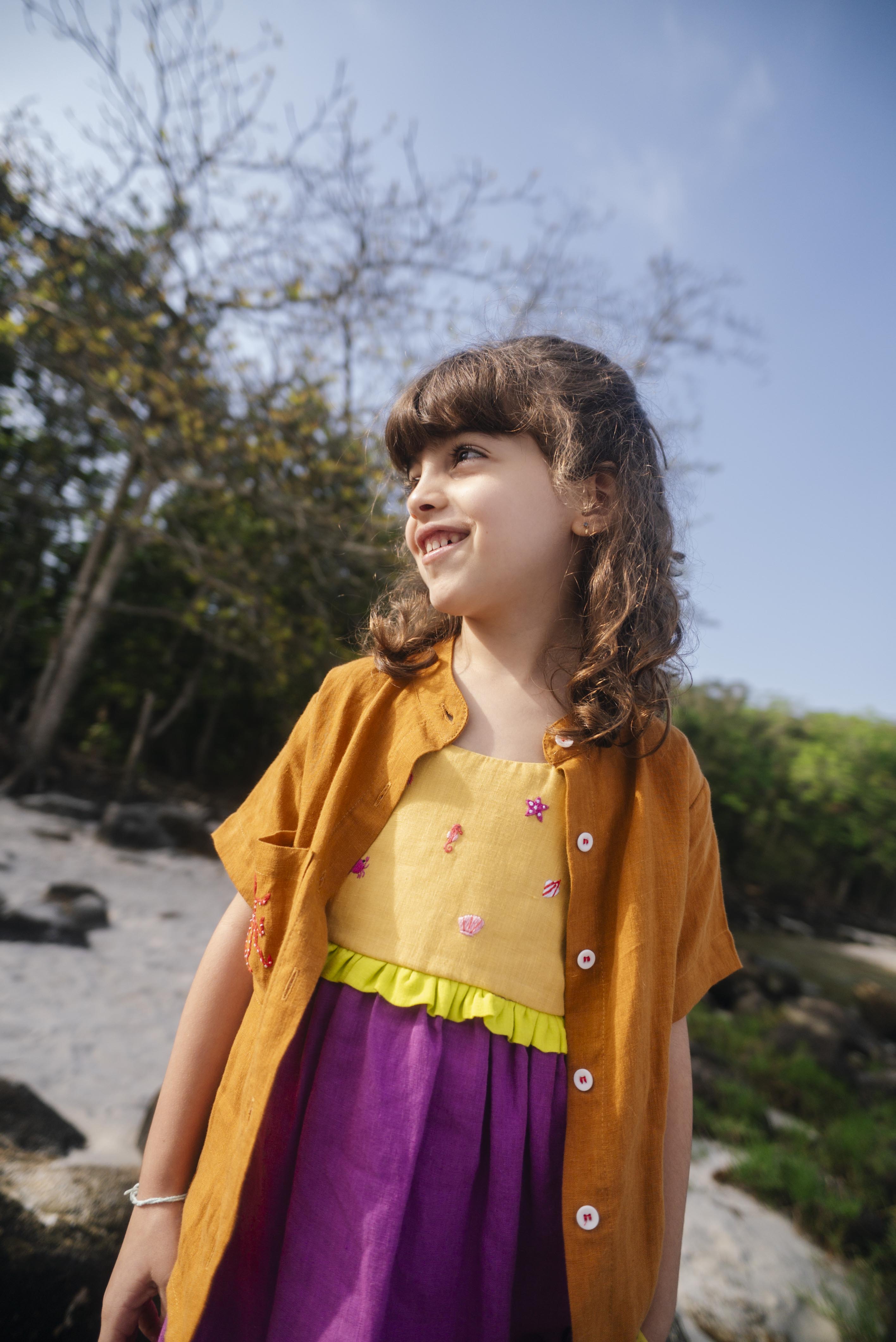 Menina usando vestido infantil em linho roxo e amarelo com bordados manuais de conchas, polvo e cavalo-marinho, com camisa marrom sobreposta.