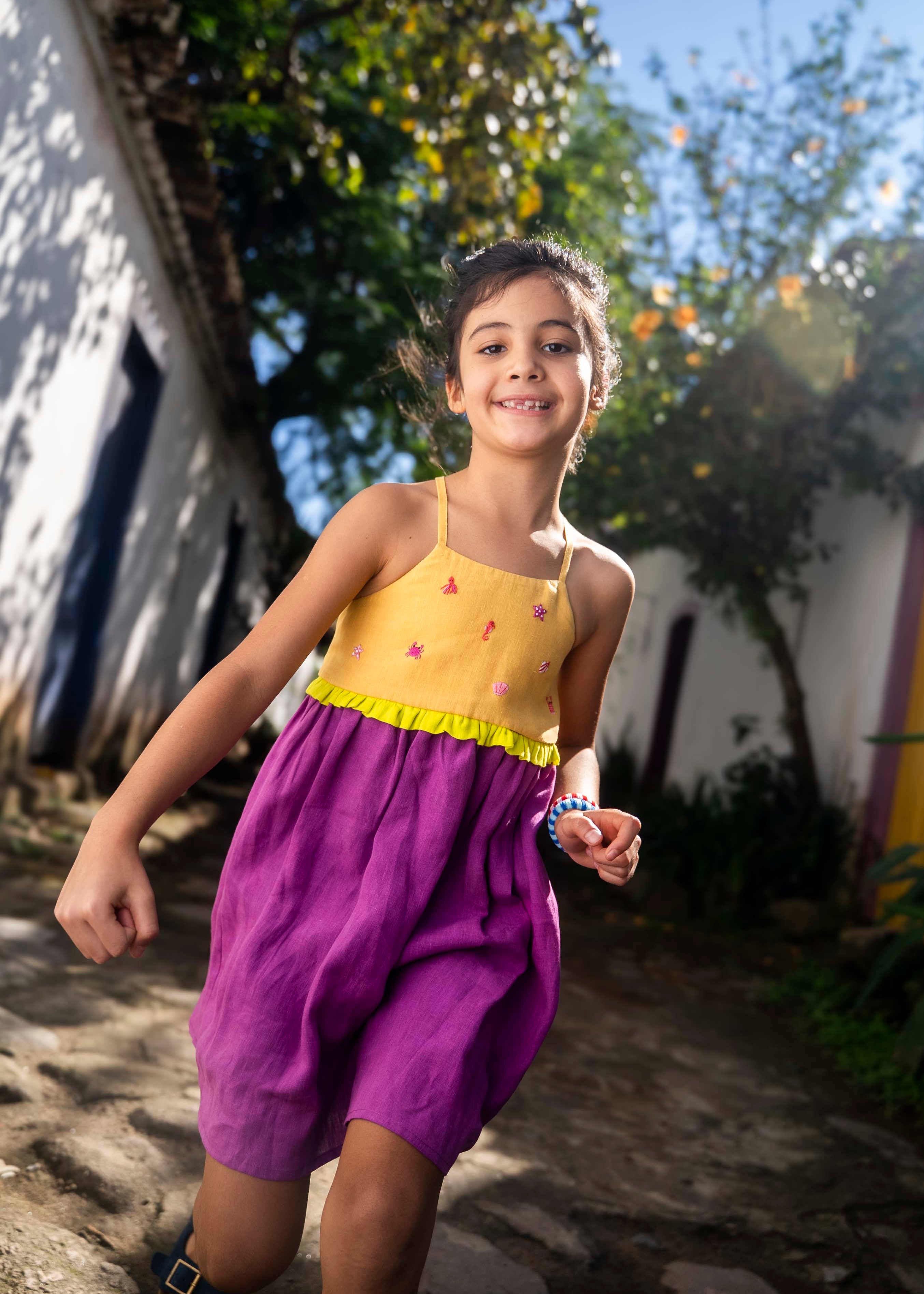 Menina usando vestido infantil em linho roxo e amarelo com bordados manuais na pala de conchas, estrelas do mar, polvo e cavalo-marinho.