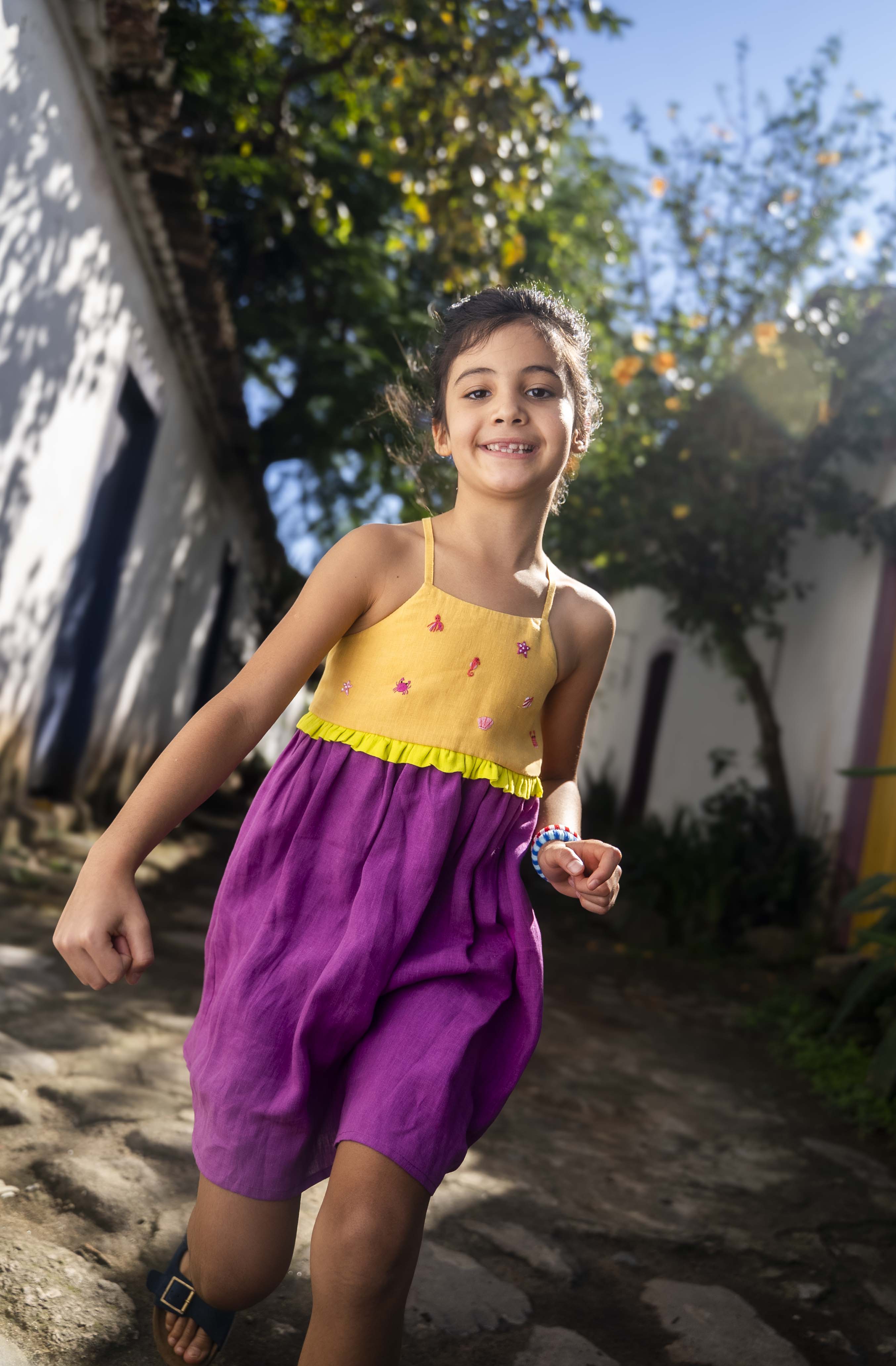Menina usando vestido infantil em linho roxo e amarelo com bordados manuais na pala de conchas, estrelas do mar, polvo e cavalo-marinho.