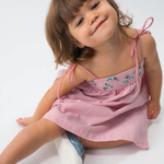 Menina sentada vestindo Vestido Flora em algodão anarruga vermelho listrado, com pala bordada e saia ampla.