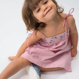 Menina sentada vestindo Vestido Flora em algodão anarruga vermelho listrado, com pala bordada e saia ampla.