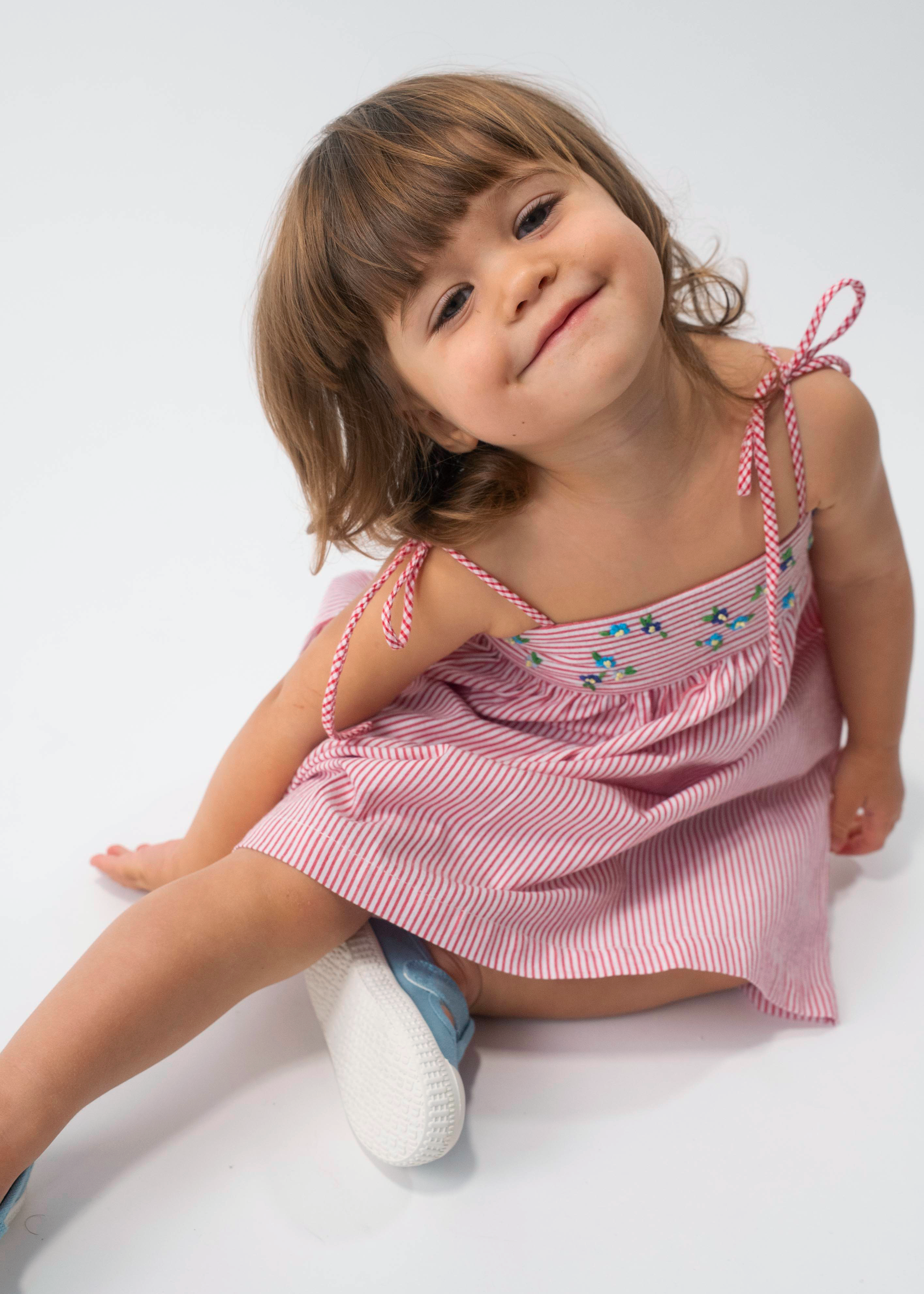Menina sentada vestindo Vestido Flora em algodão anarruga vermelho listrado, com pala bordada e saia ampla.