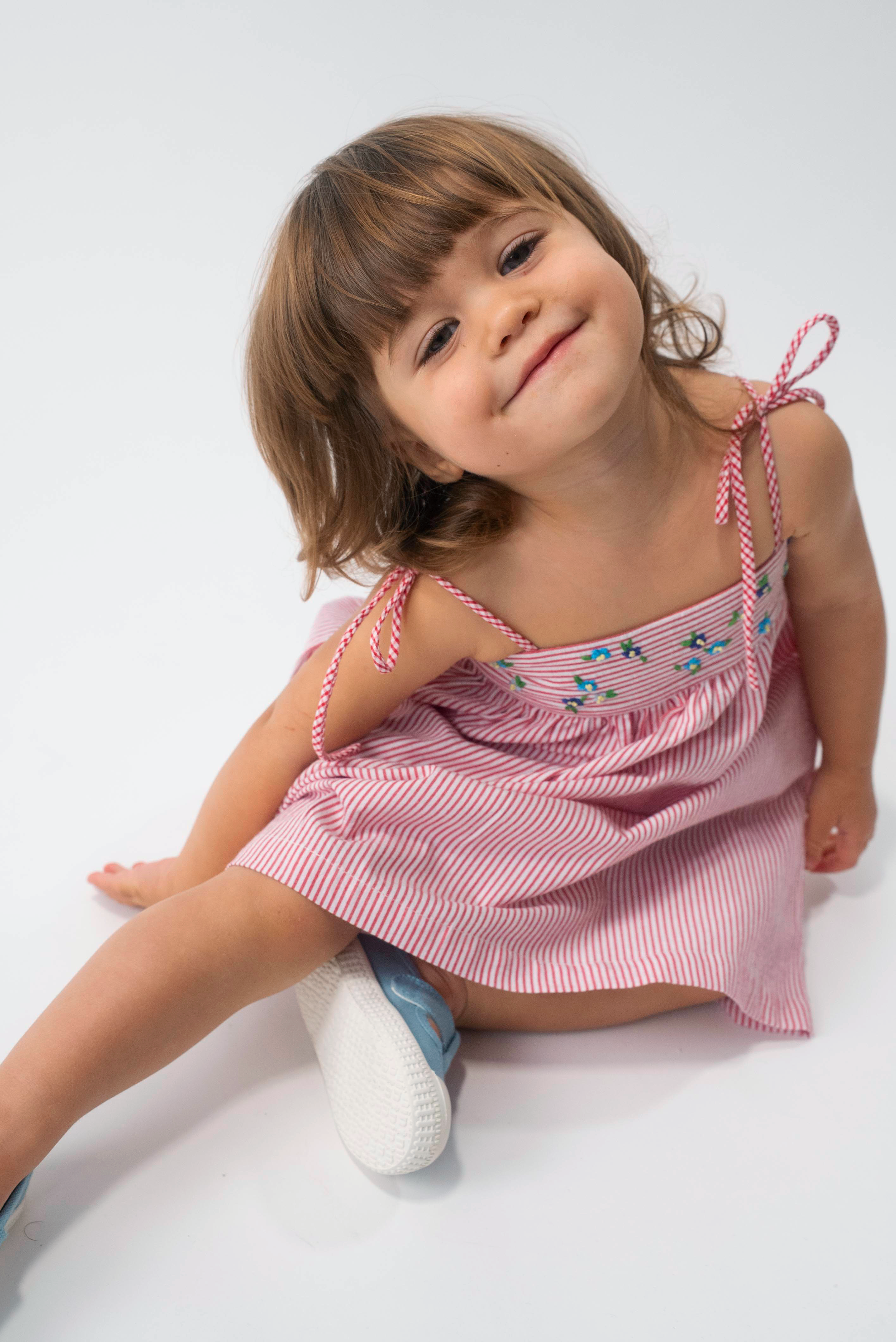 Menina sentada vestindo Vestido Flora em algodão anarruga vermelho listrado, com pala bordada e saia ampla.