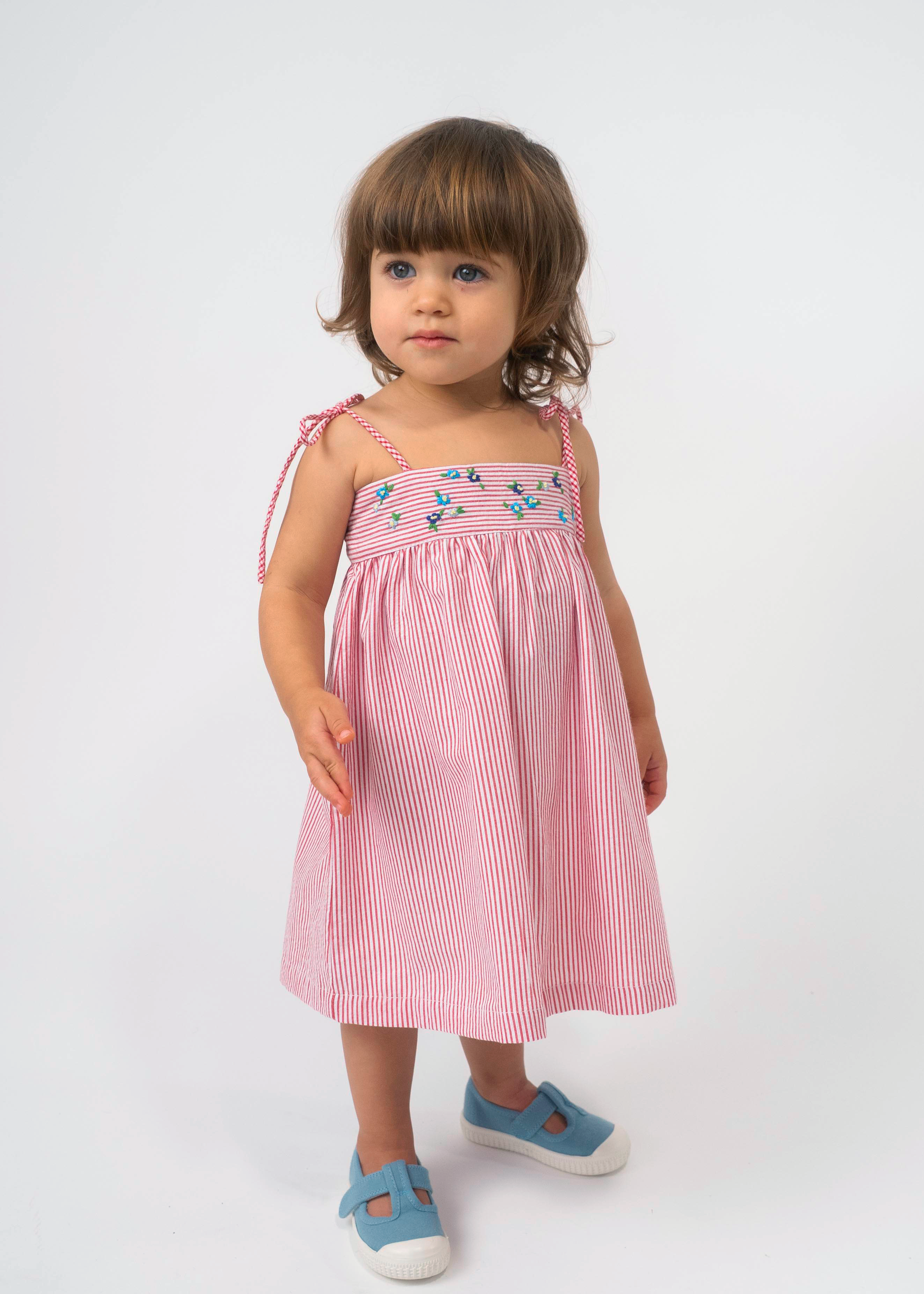 Menina vestindo Vestido Flora vermelho listrado, com flores bordadas à mão e alças de amarrar nos ombros.