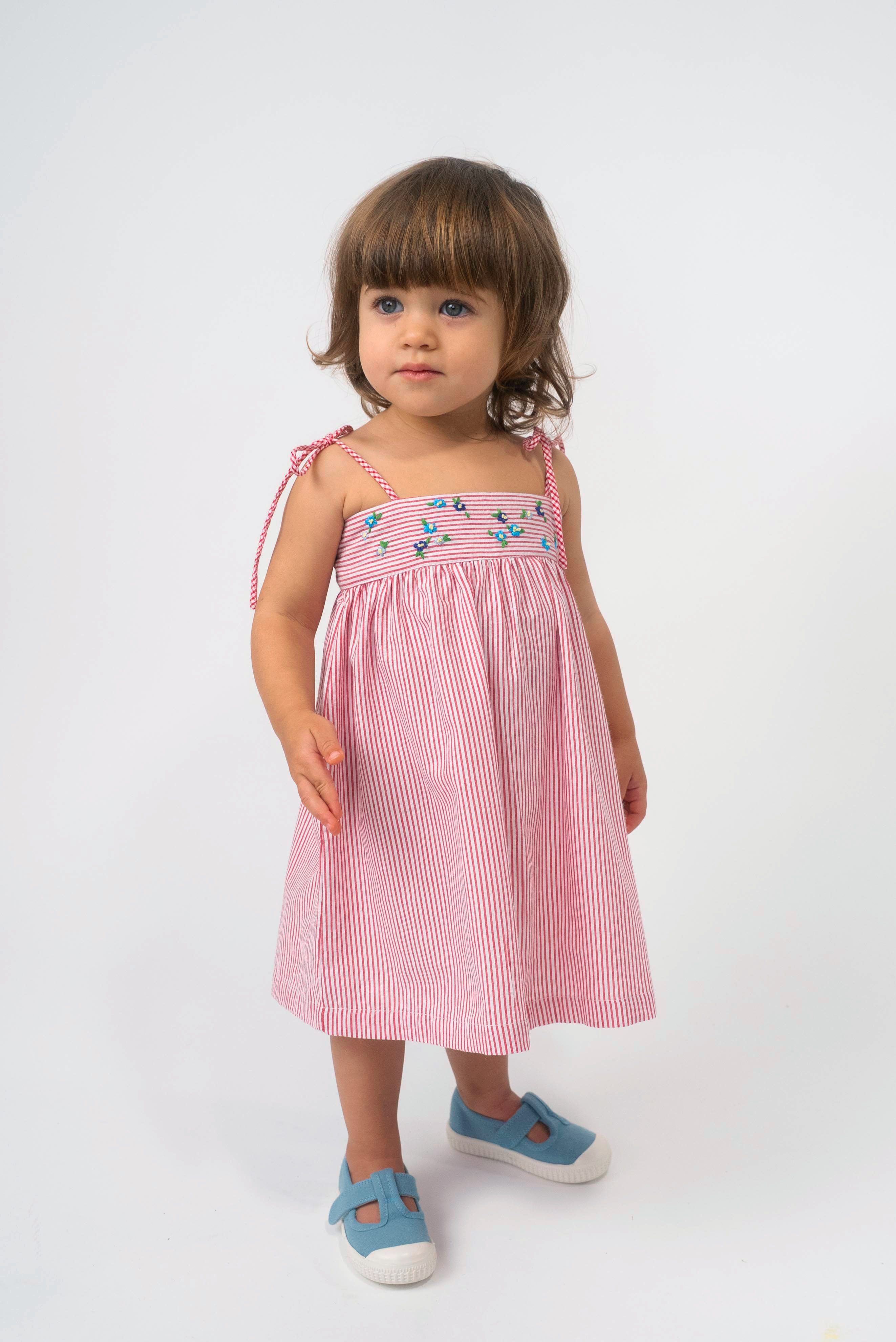 Menina vestindo Vestido Flora vermelho listrado, com flores bordadas à mão e alças de amarrar nos ombros.