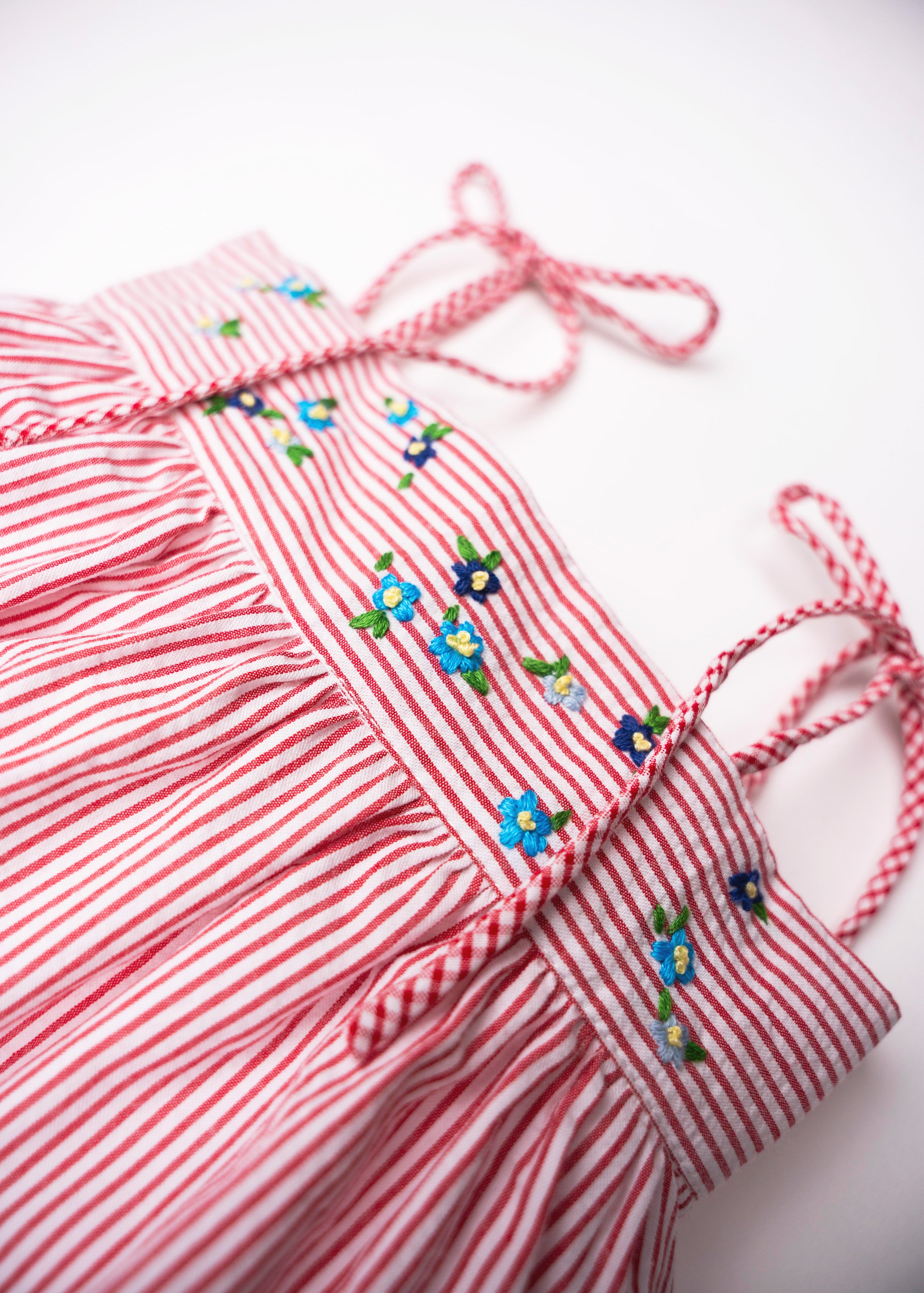 Detalhe das flores azuis bordadas à mão e das alças com efeito xadrez no Vestido Flora vermelho listrado.