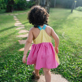 Detalhe das costas cavadas do vestido infantil em viscose Ecovero™ com alças retas azul turquesa, pala amarela e saia franzida em rosa pink. 