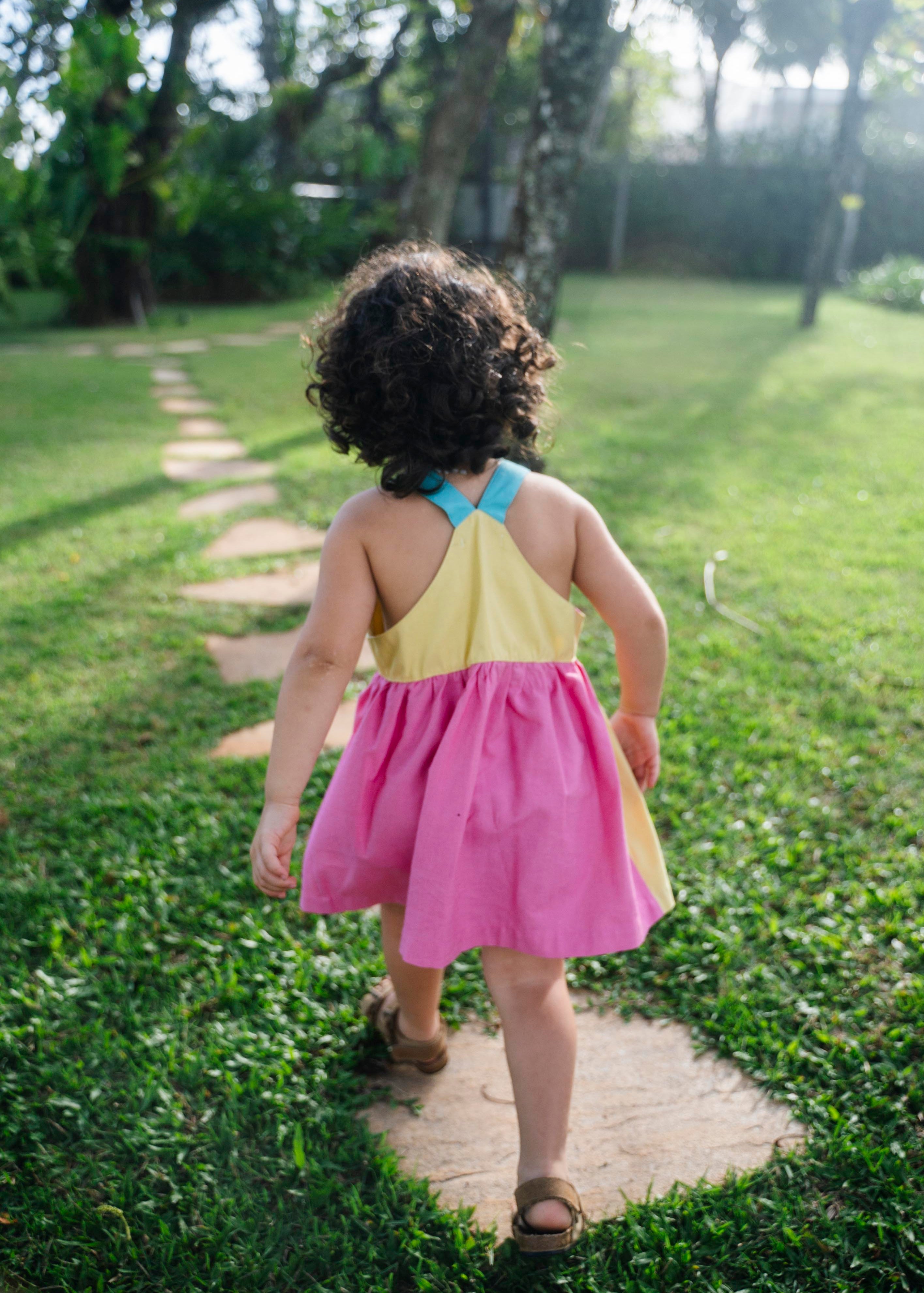 Detalhe das costas cavadas do vestido infantil em viscose Ecovero™ com alças retas azul turquesa, pala amarela e saia franzida em rosa pink. 